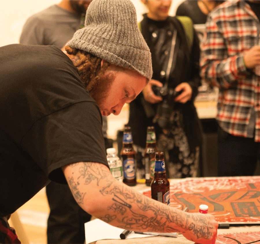 Person with tattoos and a beanie at a table with beer bottles and a red and white checkered tablecloth.