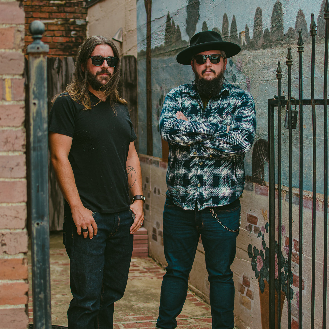 Image of Sons of Gulliver - a ONE on Stage Band. Two people standing in alleyway. Landscape mural on wall behind them.