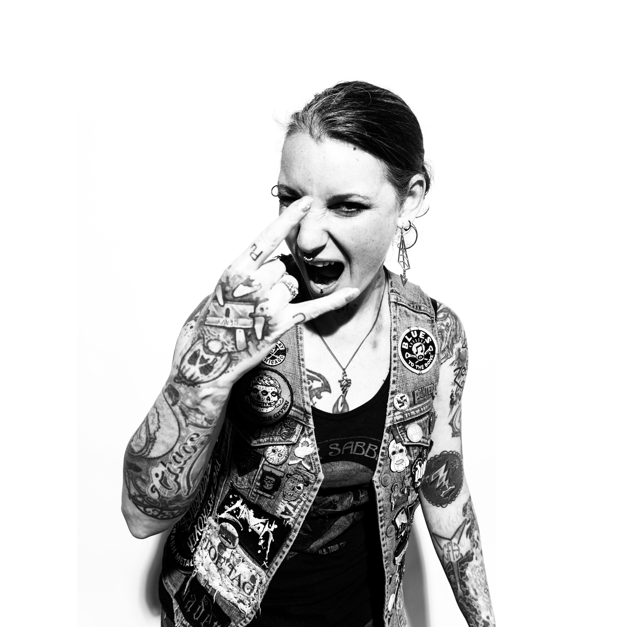 B&W photo of person holding up a "ROCK ON" hand symbol. Person has tattoos, large earrings, and a patterned vest. 