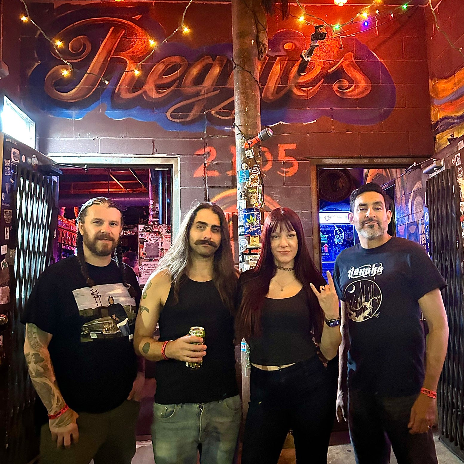 Image of Black Road - a ONE on Stage band. Four members standing in venue. Wall in background says "REGGIE'S."
