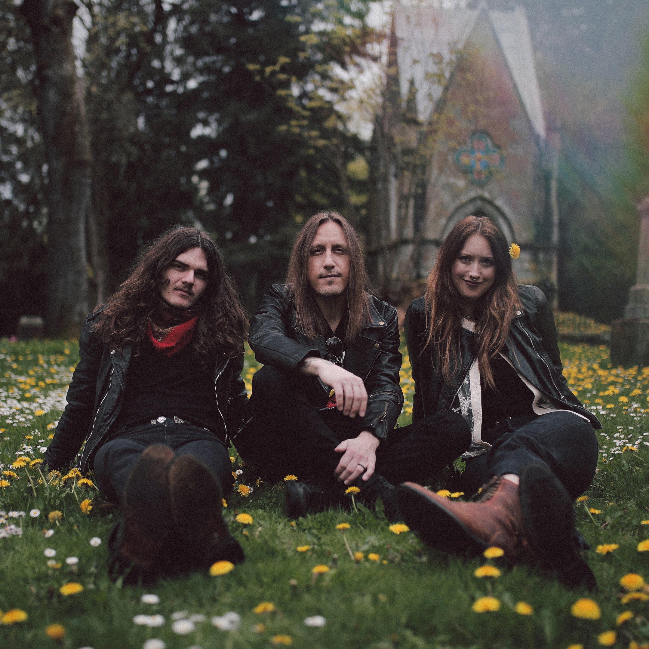 Image of Hippie Death Cult - a new ONE On Stage band. Image shows three people (band members) sitting in a bed of grass dotted by dandelions. All band members are wearing black. A church-looking stone building and trees are behind them.